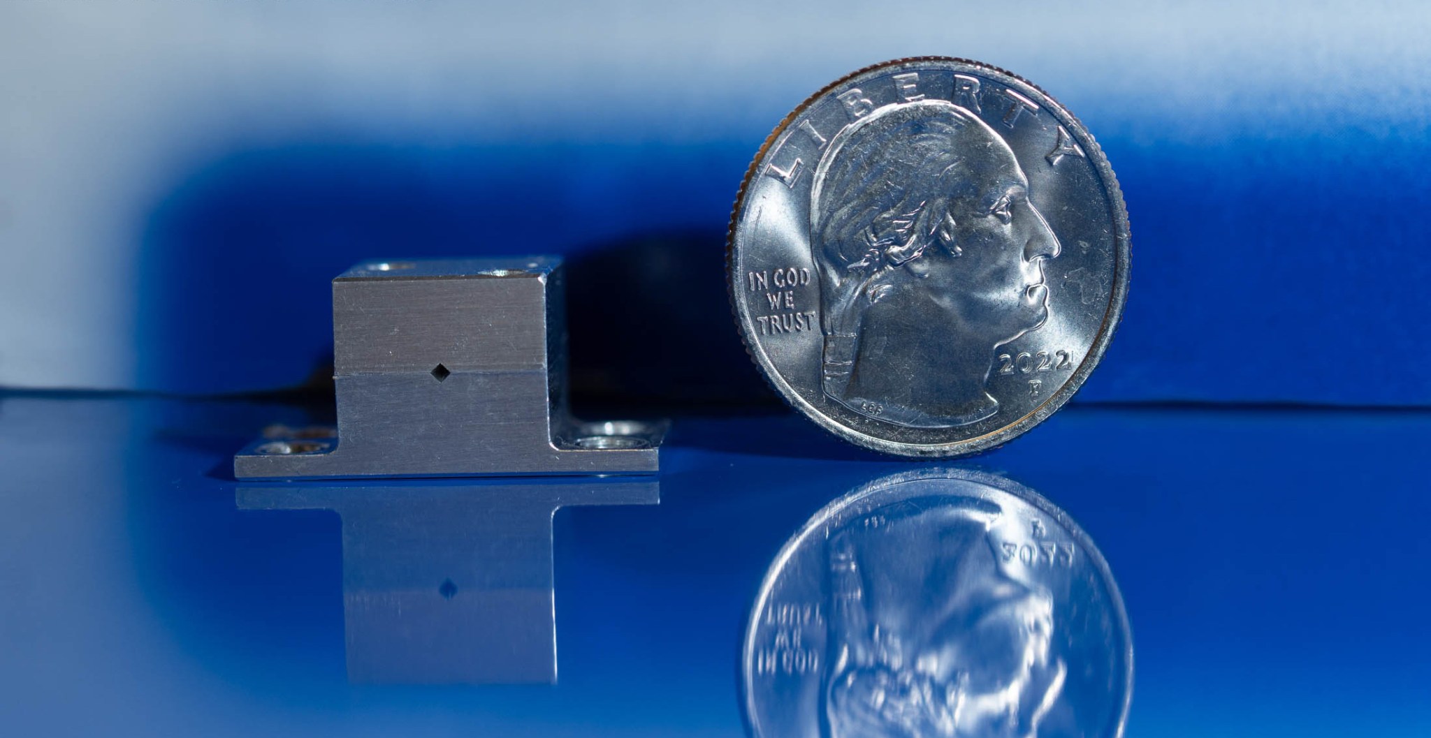 a small metal block sits next to a much larger quarter, reflected on a shiny surface with a blue background