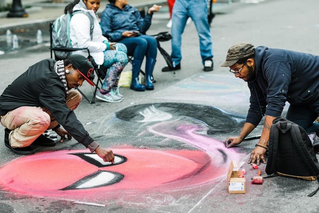 Two men using sidewalk chalk to draw a spiderman and venom head on the ground.