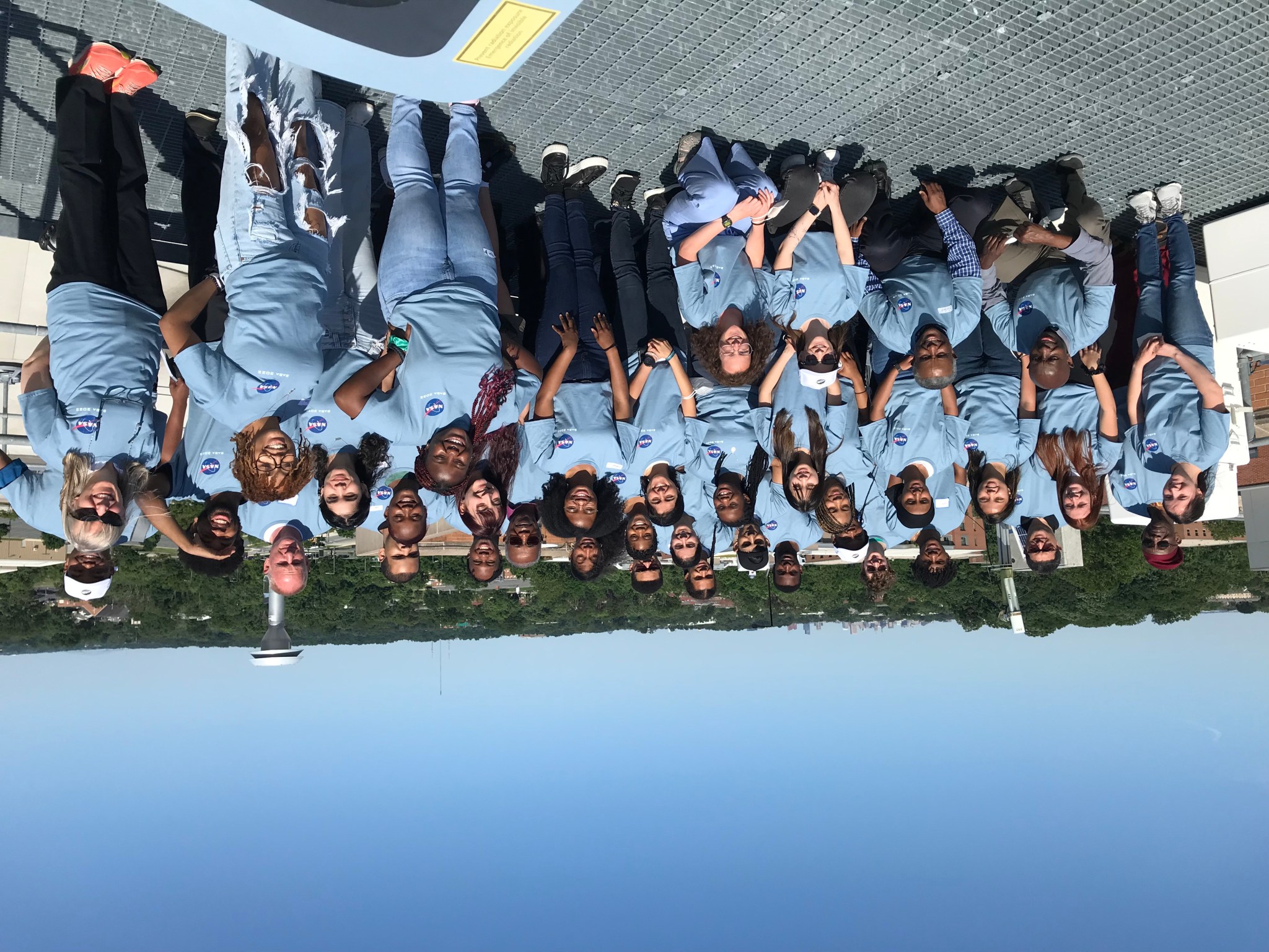 A large group of people, all wearing light blue t-shirts with the NASA logo, pose on a rooftop.