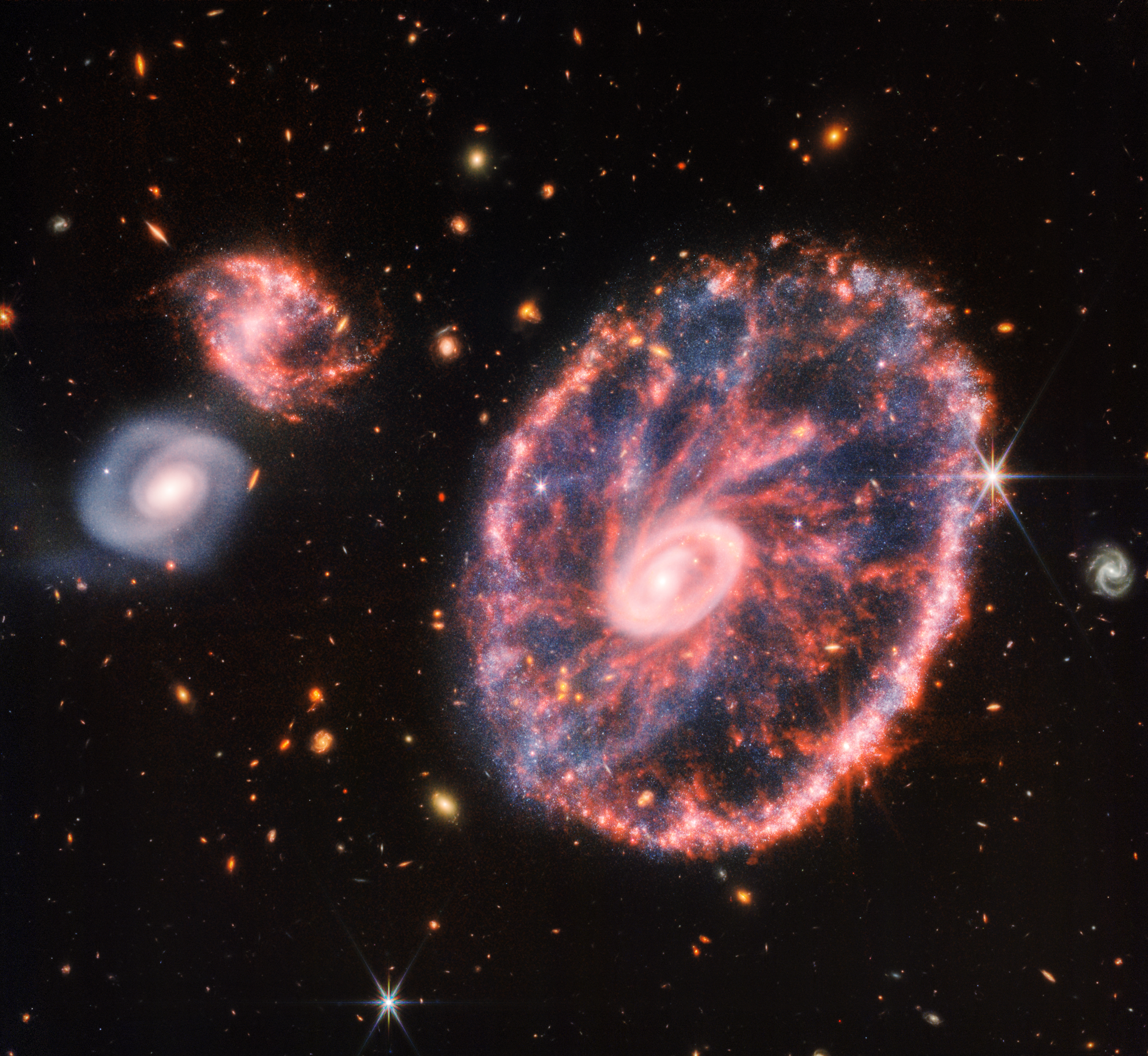 A large pink, speckled galaxy resembling a wheel with with a small, inner oval, with dusty blue in between on the right. Two smaller spiral galaxies about the same size are to the left, all against a black background.