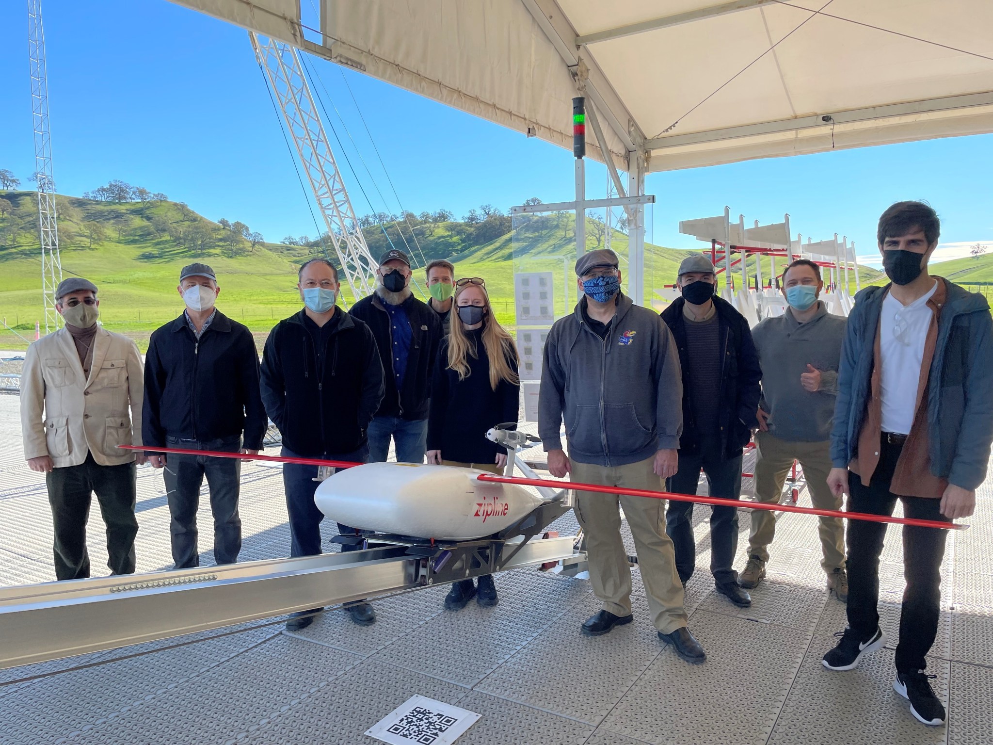 Group photo of NASA researchers at the Zipline Test Facility with a large vehicle model in front of them.