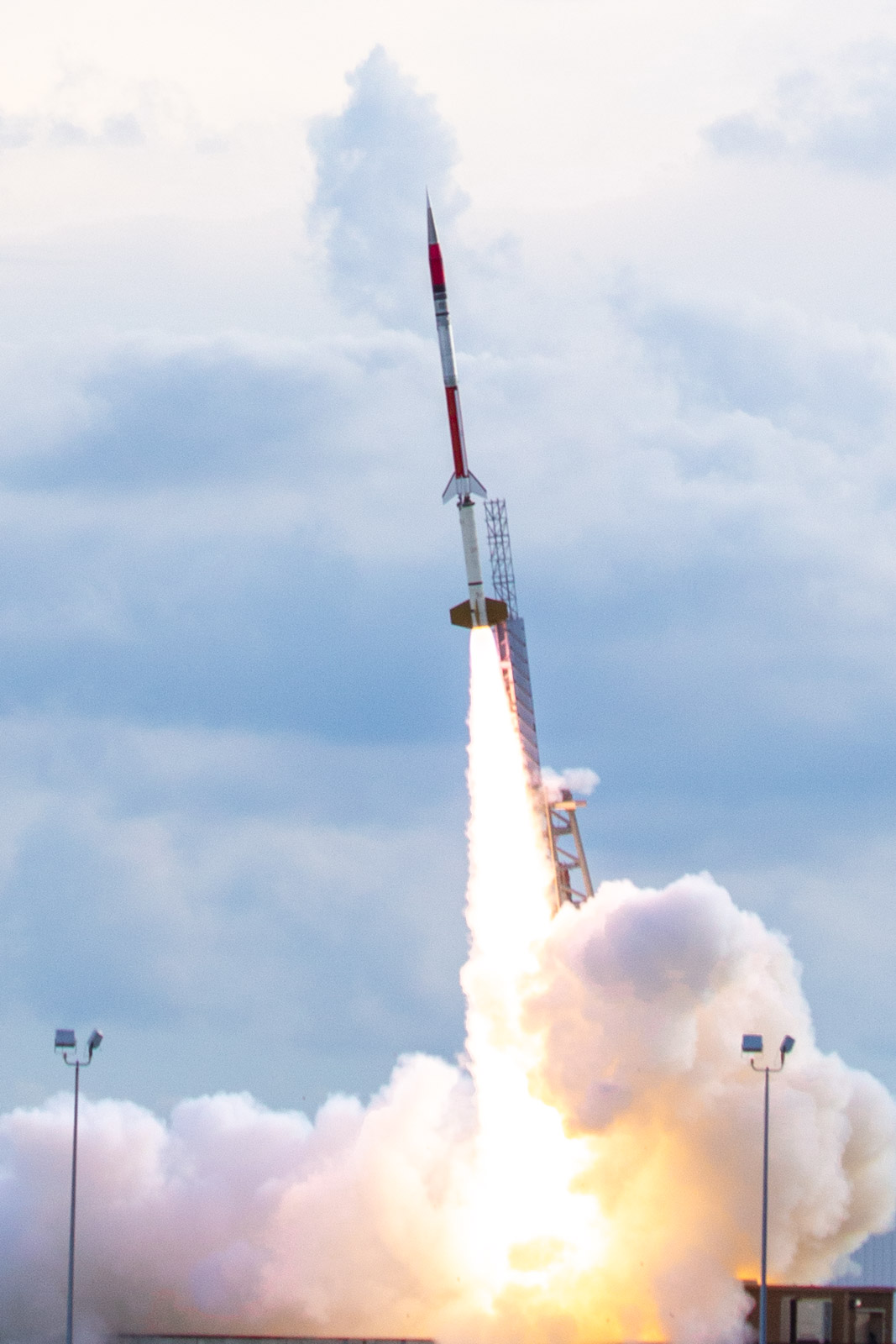 A red and silver small rocket in flight, launching just off its launch pad with a plume of fire and smoke underneath.
