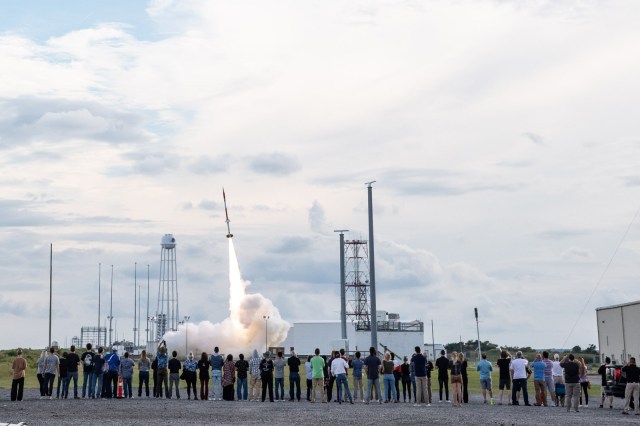 In the foreground, a line of people stand at a safe distance away from a sounding rocket launching in the background. The rocket is just off the pad with a plume of fire and smoke underneath.