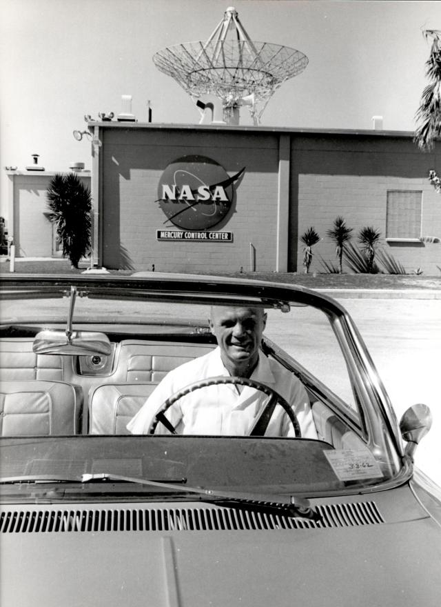 Astronaut John H. Glenn sits in a convertible car in front of the east side of the Mercury Mission Control building.