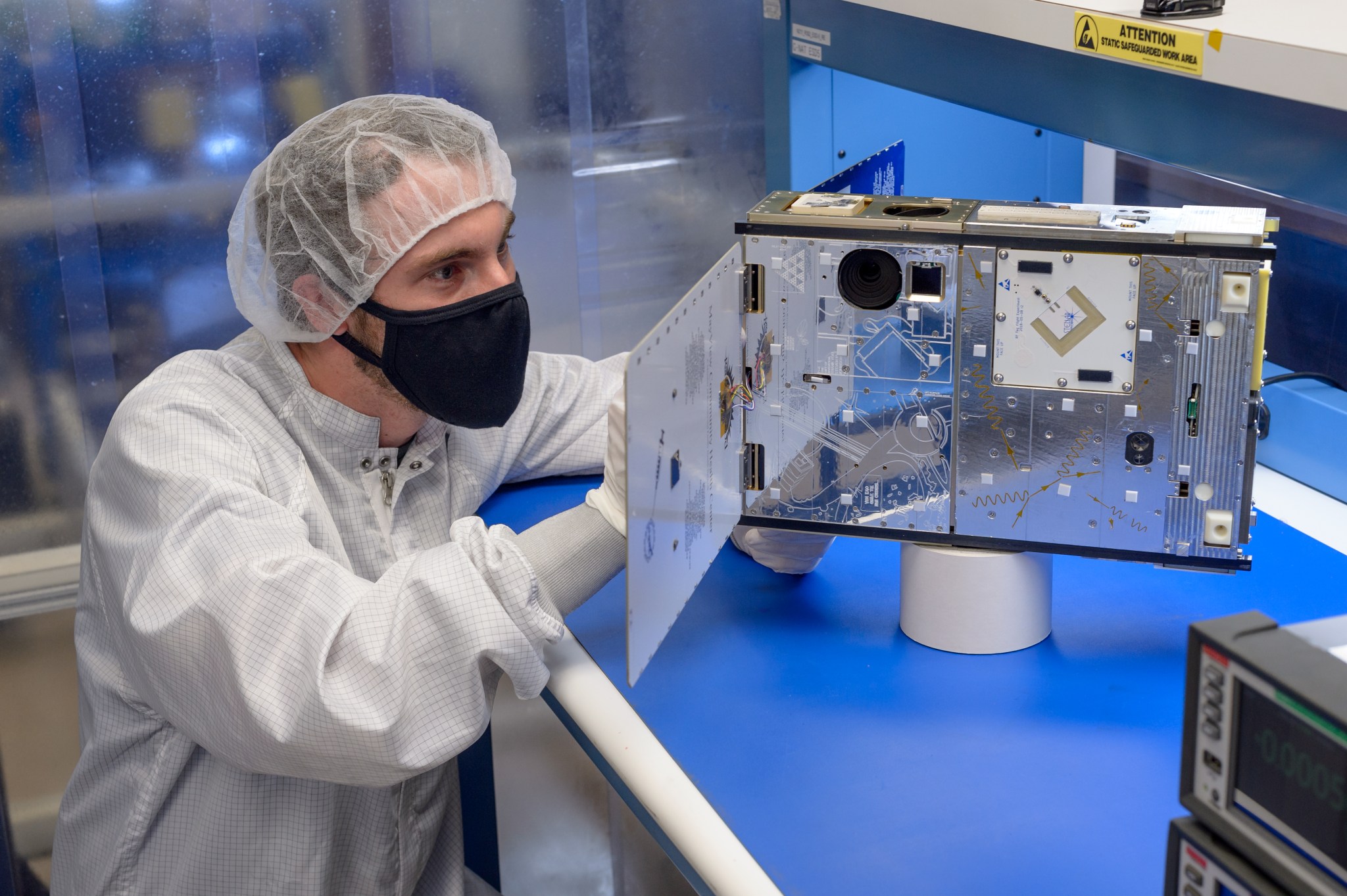 A person in a white clean-suit and hair net and black mask is crouched down inspecting a reflective, silver CubeSat device with two panels extended from the sides.