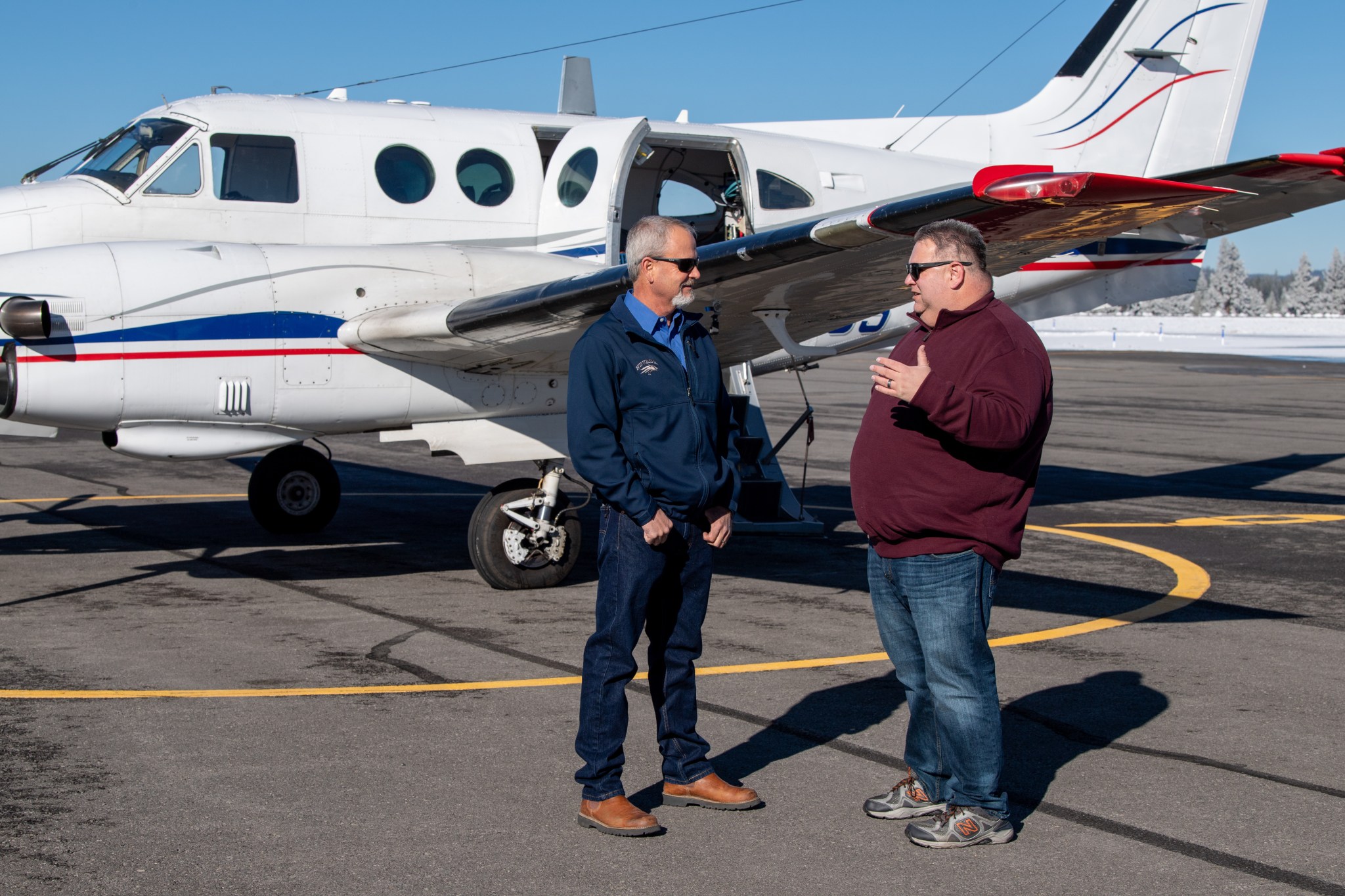 Photo of two white men standing in front of a plane talking to each other.