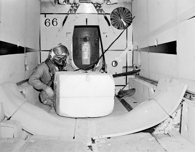 Man with flight suit and helmet watches a package in bomb bay of a military aircraft.