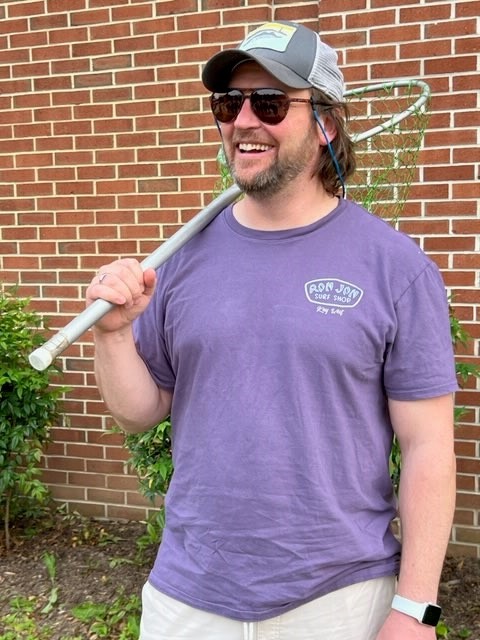 A man in a purple T-shirt holds a fishing net over one shoulder and smiles.