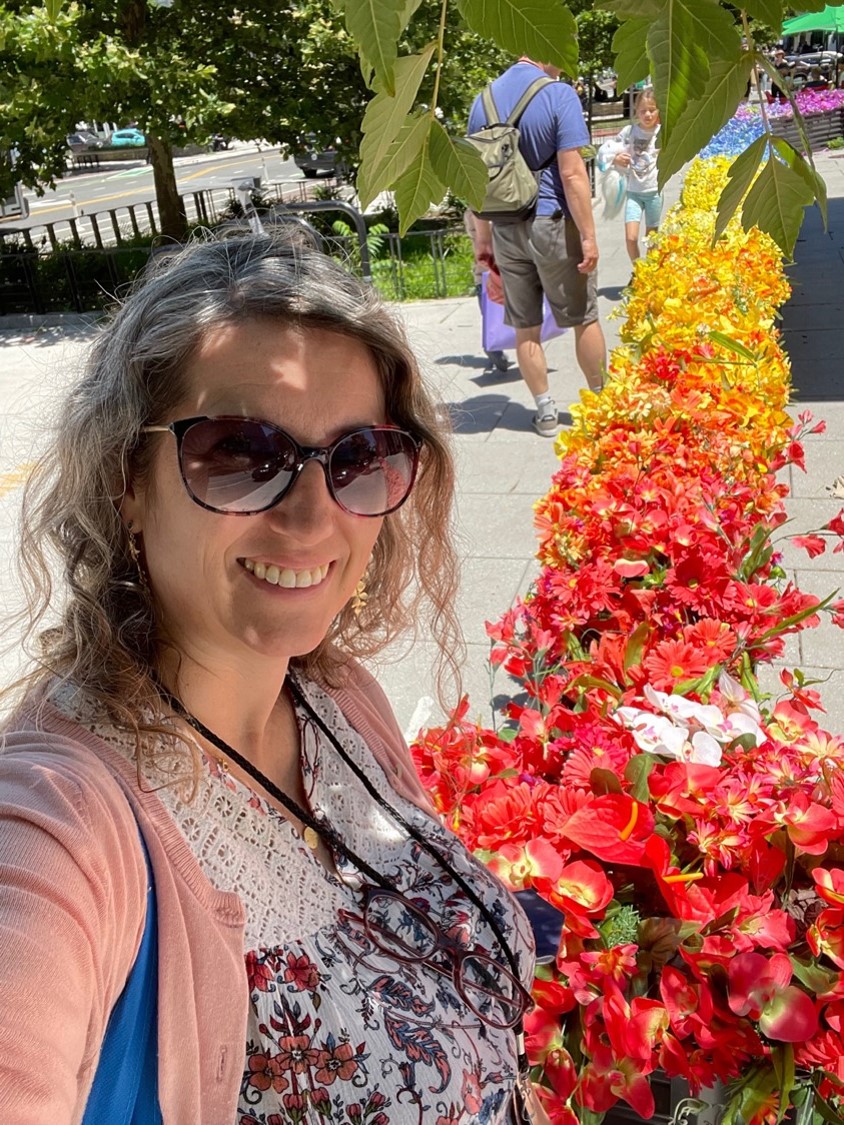 A white woman with blonde wavy hair takes a selfie in front of a rainbow-colored row of flowers.