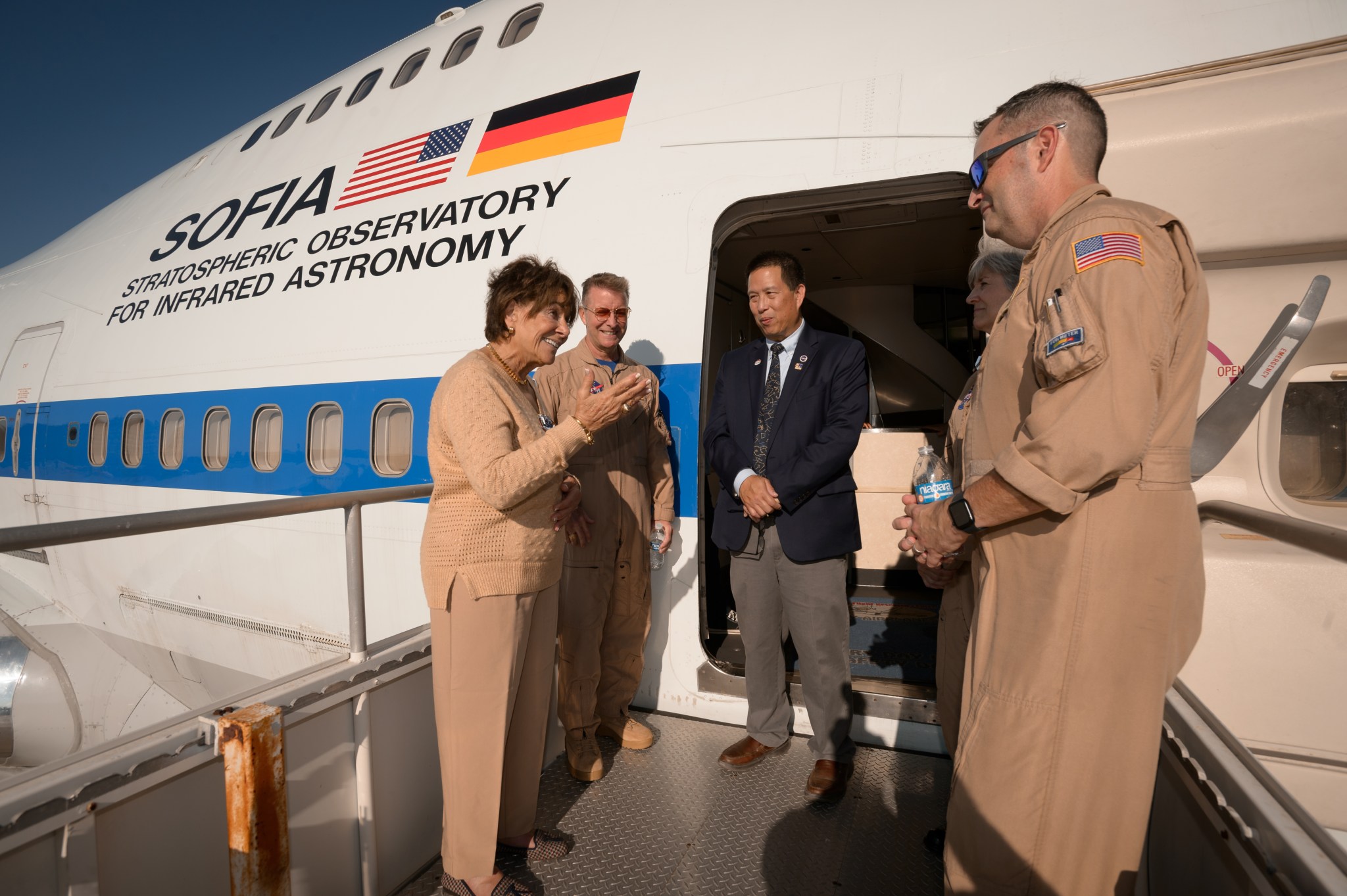 A group of five people, several in brown flight suits, stand on the stairs outside an aircraft. "SOFIA Stratospheric Observatory for Infrared Astronomy" is written on the side of the plane.