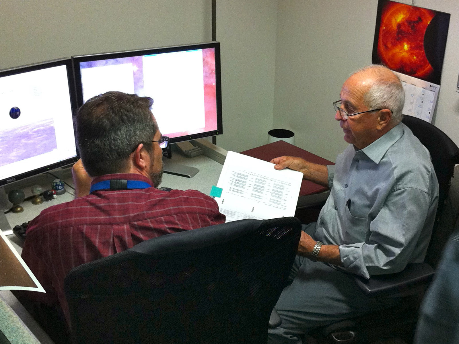 Two men sit at a desk, facing a computer screen. The man on the right holds a binder.