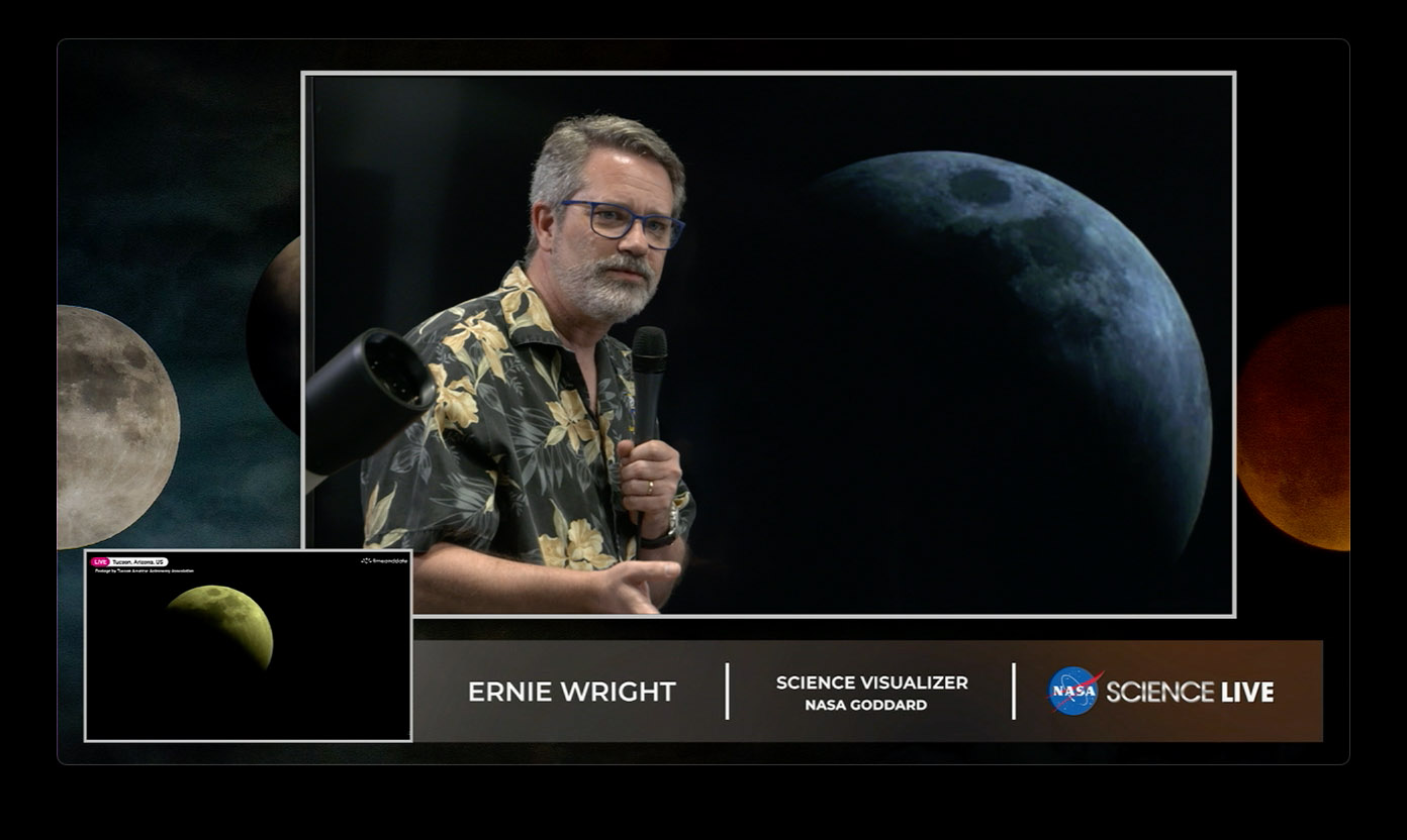 A man with gray hair, beard, and glasses speaks into a microphone in front of a large image of the Moon.