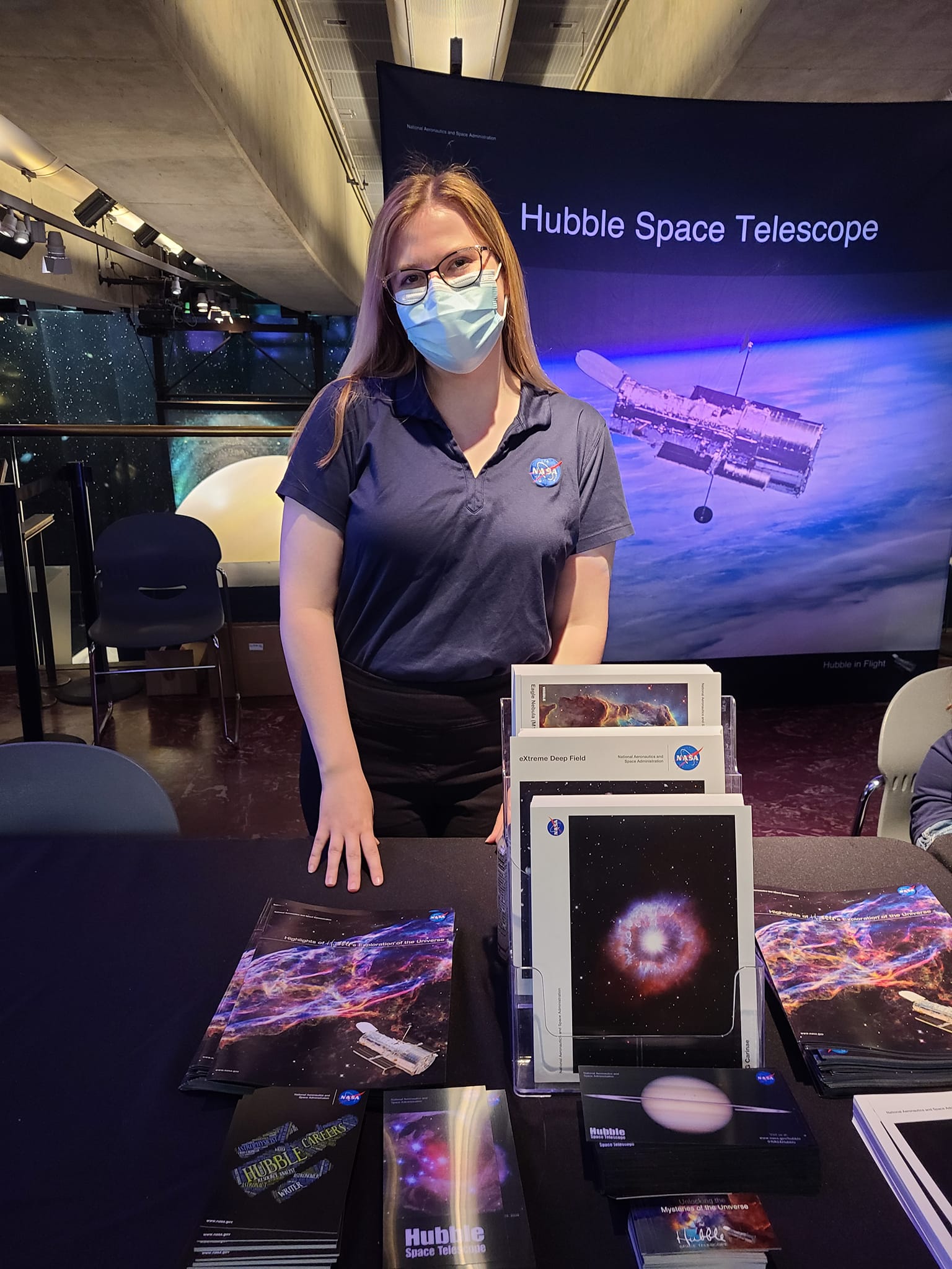 Elizabeth Tammi, a woman with glasses and long blonde hair, stands at a table with printed Hubble flyers.