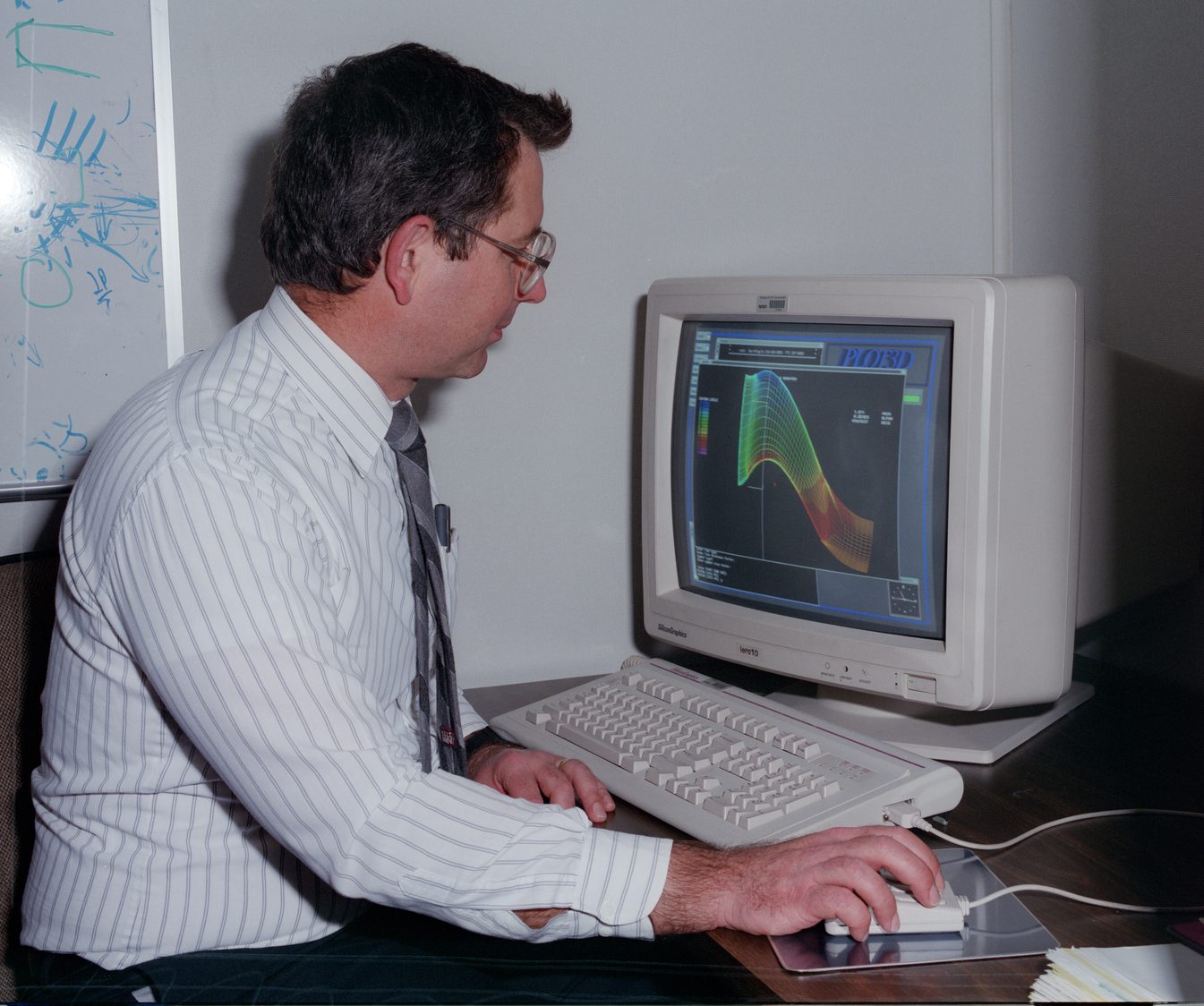 Older photo of a man sitting at a 1990s era computer with a colored graph image on the monitor. In the background is a whiteboard with blue mathematical scribbling.