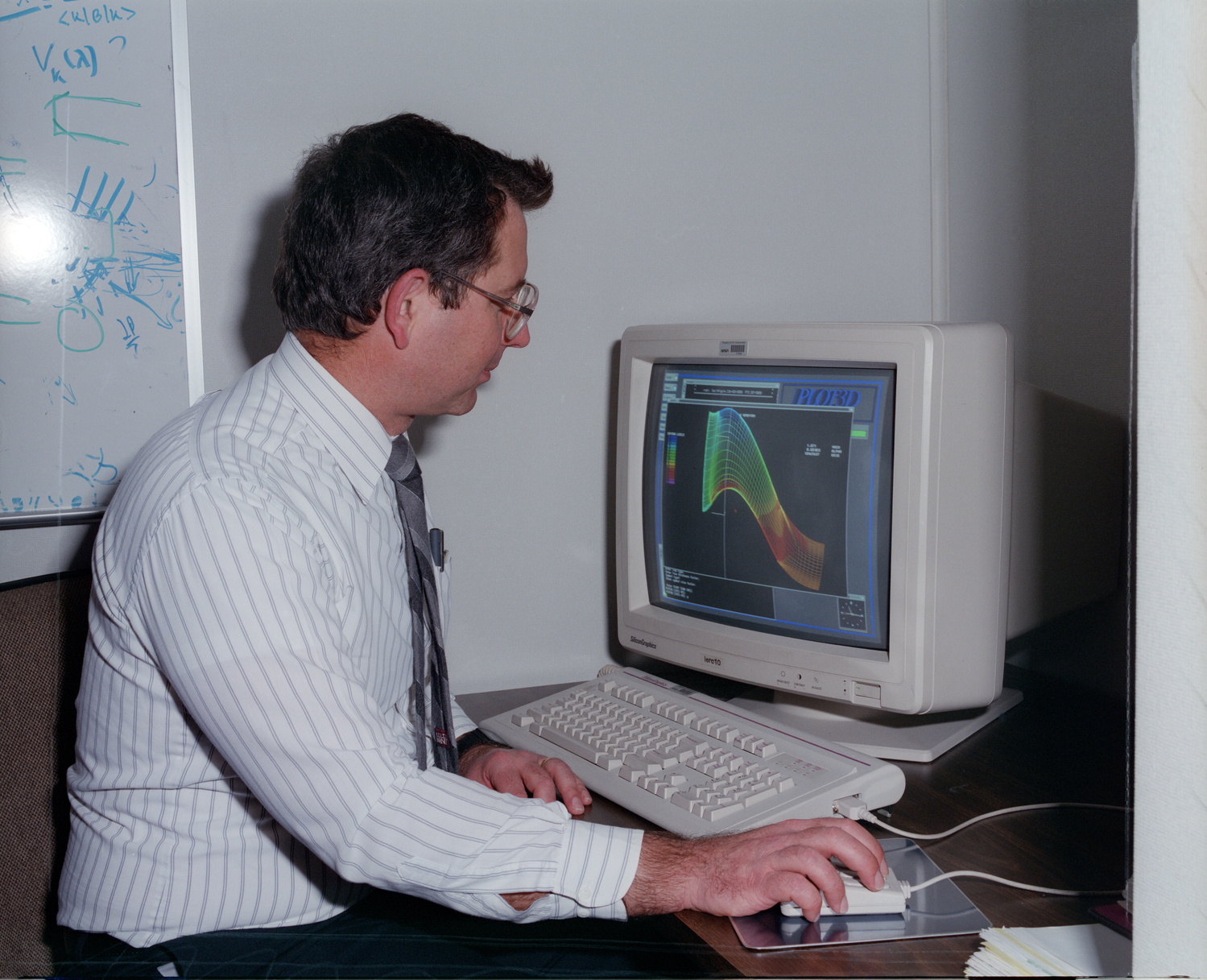 Older photo of a man sitting at a 1990s era computer with a colored graph image on the monitor. In the background is a whiteboard with blue mathematical scribbling.
