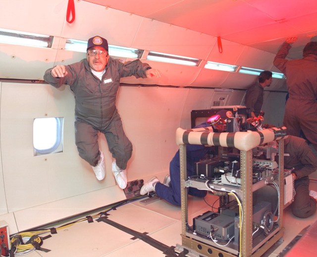 Man in flight suit wearing cap, floats inside an aircraft near a rig holding an experiment.