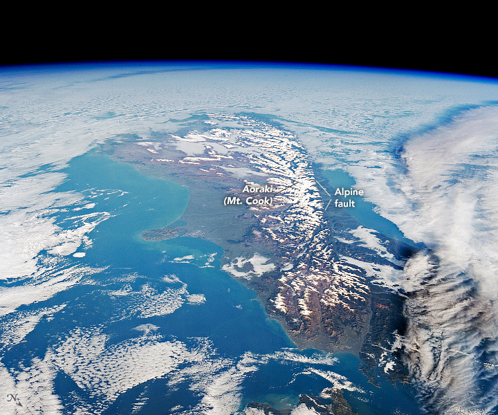 Photo of New Zealand's South Island as viewed from the International Space Station. The island is shades of dark brown, with a snow capped mountain range running along its center. The surrounding ocean is bright blue.