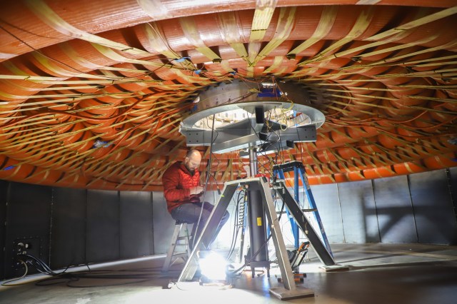 A person standing underneath a disk-like cone structure with straps connecting a set of circular tubes. A silver metal stand props the device up, and the person under sits on a small ladder examining wiring.