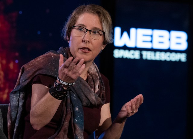 A fair-skinned woman with short light brown hair, wearing a chunky galaxy scarf, and thin-framed glasses looks off to the side, speaking with her hands. Behind her is faded text that says "WEBB SPACE TELESCOPE"