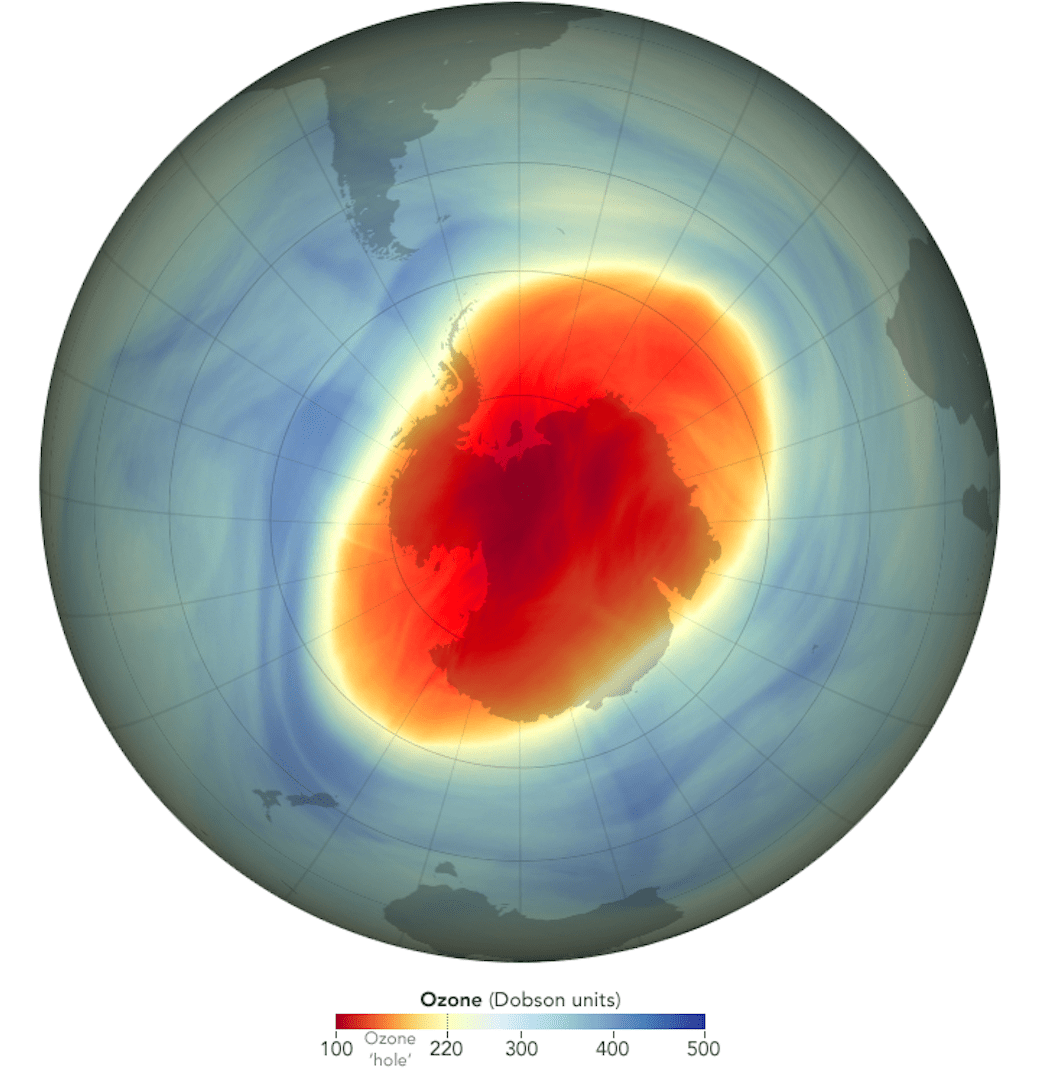 A globe centered on the South Pole, with colors indicating ozone concentration. The edges of the map are blues and greens, indicating higher ozone levels. Over Antarctica, an oval of reds and oranges shows the lower concentrations of the ozone hole.