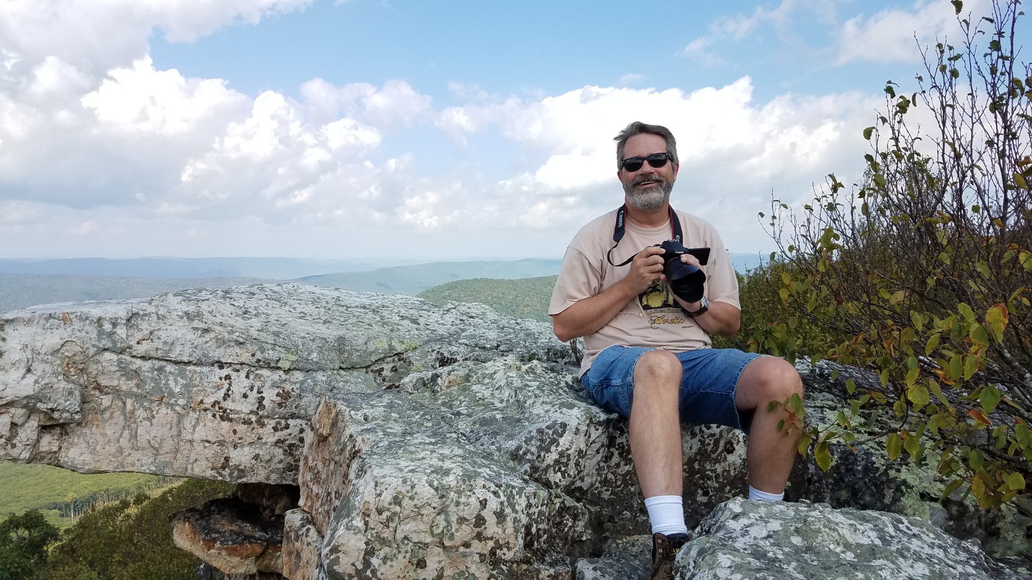 A man with gray hair, beard and sunglasses sits on a rock overlooking hills, holding a camera.