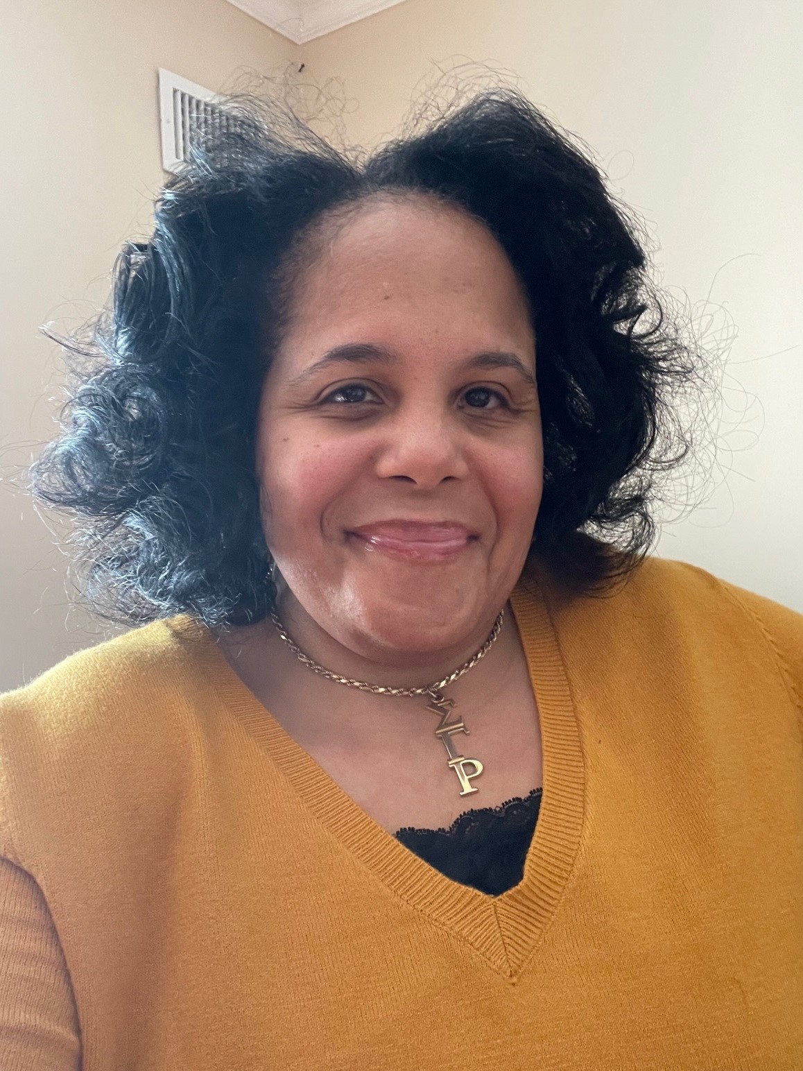 Melissa Gates, a woman with light brown skin and curly dark hair, smiles at the camera in a selfie. She wears a gold sweater and necklace.