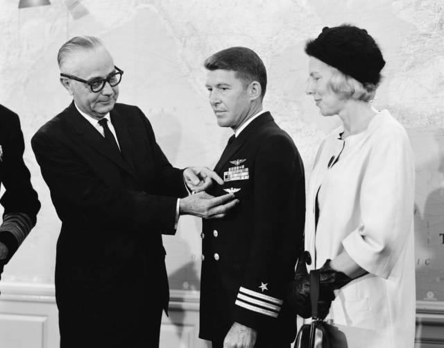Wally Schirra recieving a medal from another man with his wife looking on.