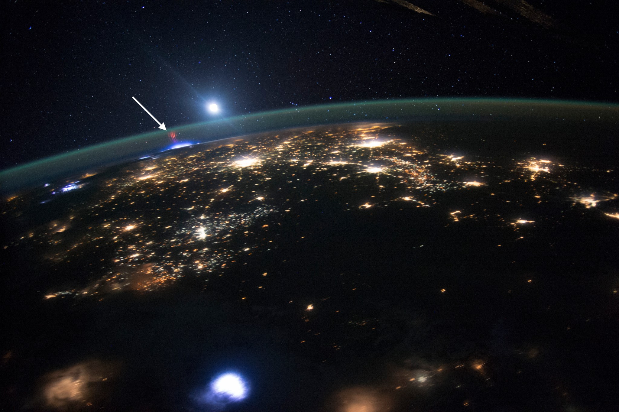 A view from Earth orbit at night. A thin, translucent green layer of the atmosphere veils clusters of city lights on Earths surface below. Over Earths limb, the blue light of a lightning storm has a small reddish light that appears above it -- a sprite.