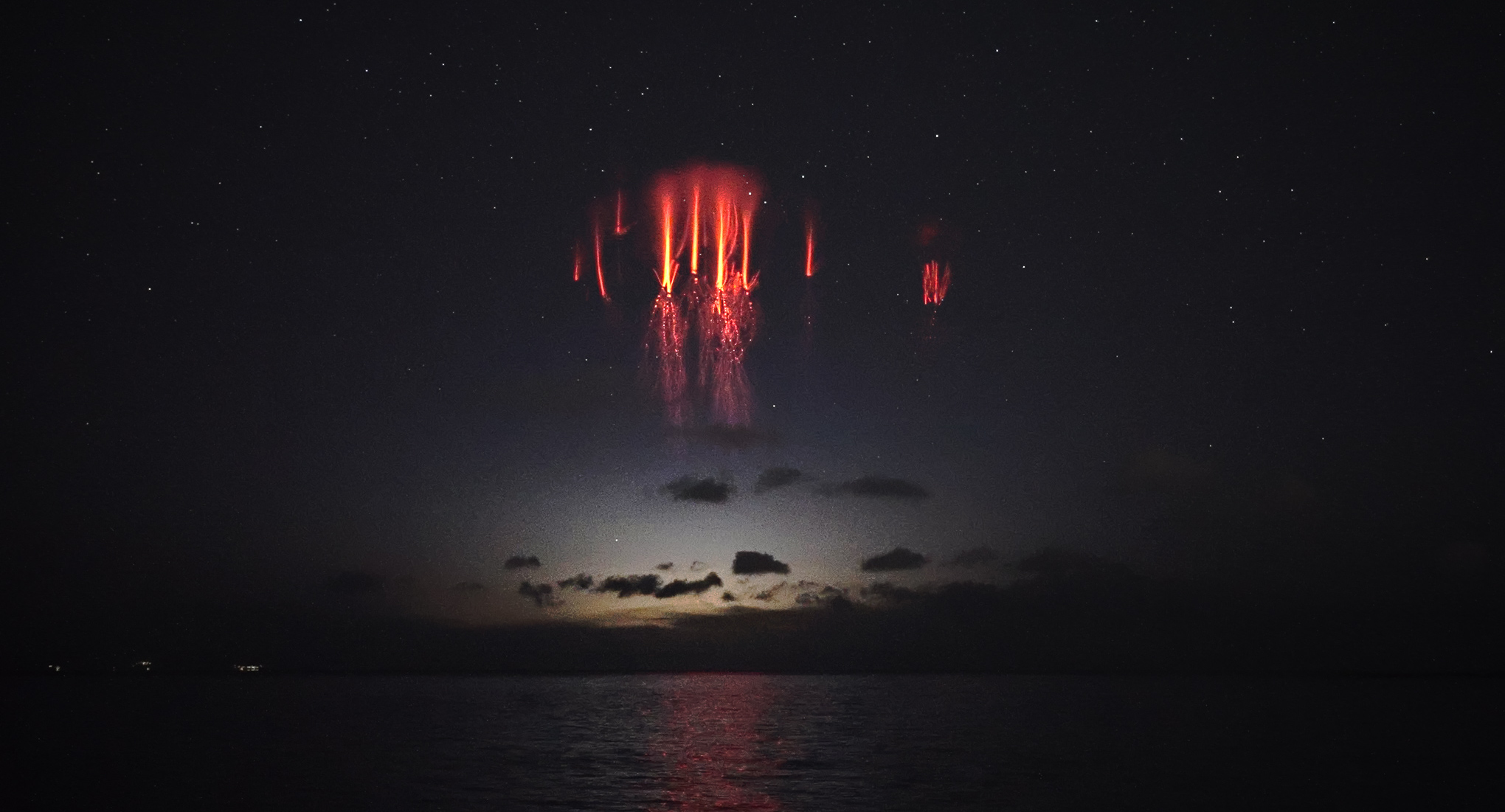 A red sprite looms over a thundercloud above the ocean. Moonlight reflects off the water.