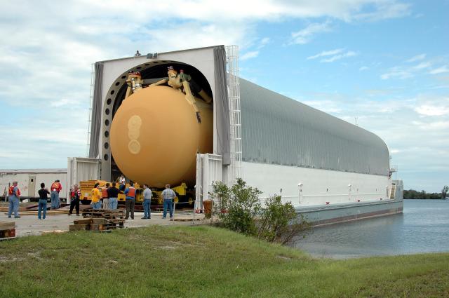 A large barge is caring a space shuttle tank that is cylindrical and orange.