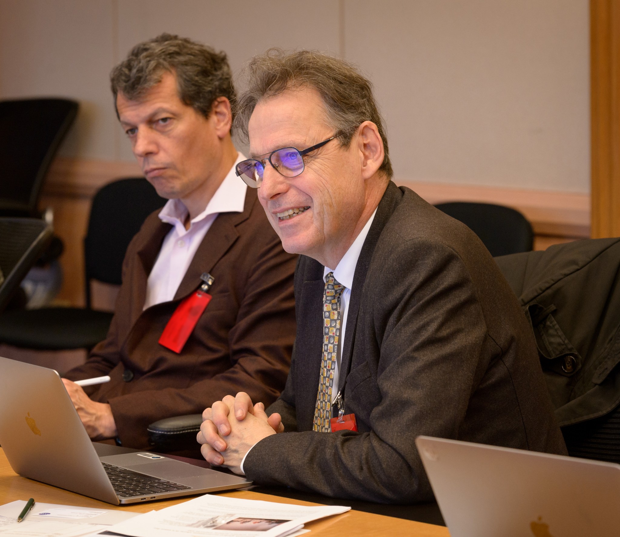 A man with black glasses wearing a suit leans forward against a table over his laptop, with another man in the background.