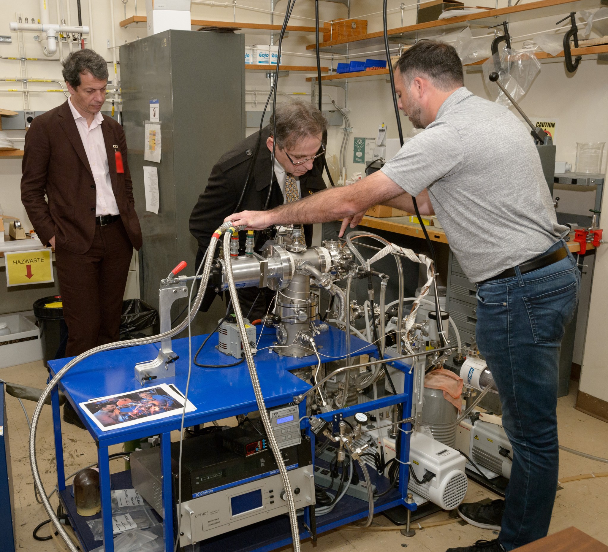 Three men in a room inspect a machine on a blue rolling cart. The machine has tubes and pipes, one man is bent over to get a close look.