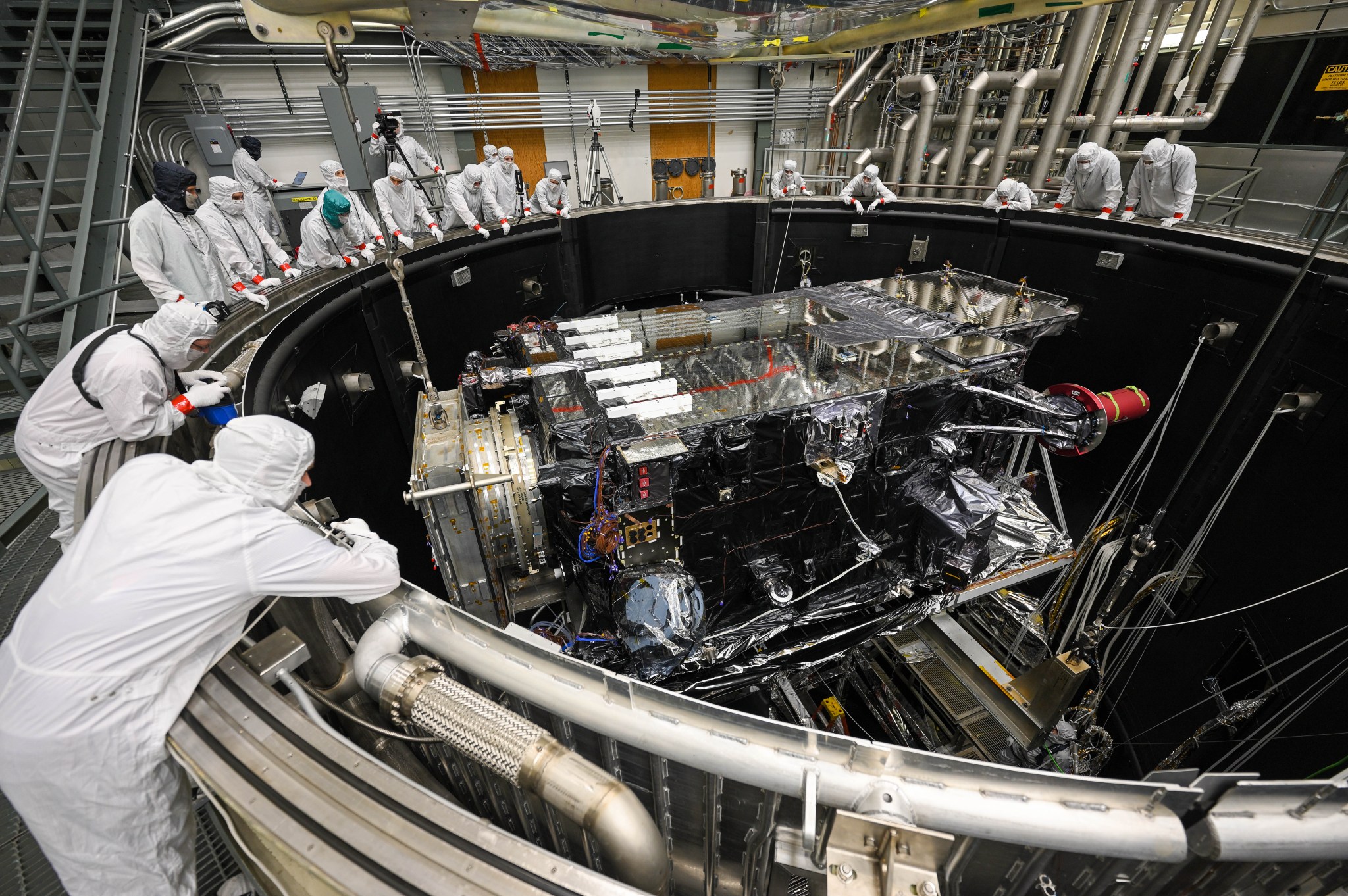 A group of people in white coveralls lean on the edge of a large cylindrical chamber, which holds a box-shaped spacecraft with many wires and panels on it.