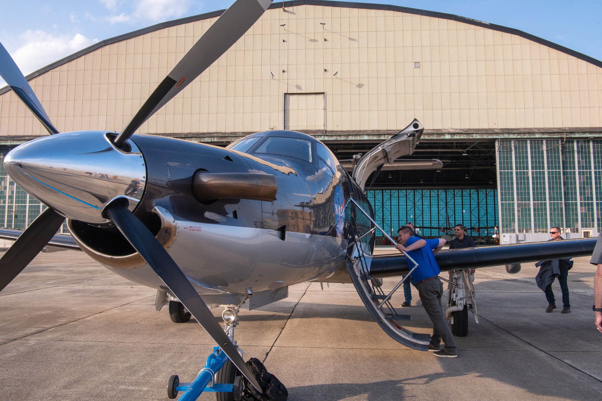 Man in blue shirt looks inside an aircraft from the drop-down steps. The aircraft sits in front of a large hangar building.
