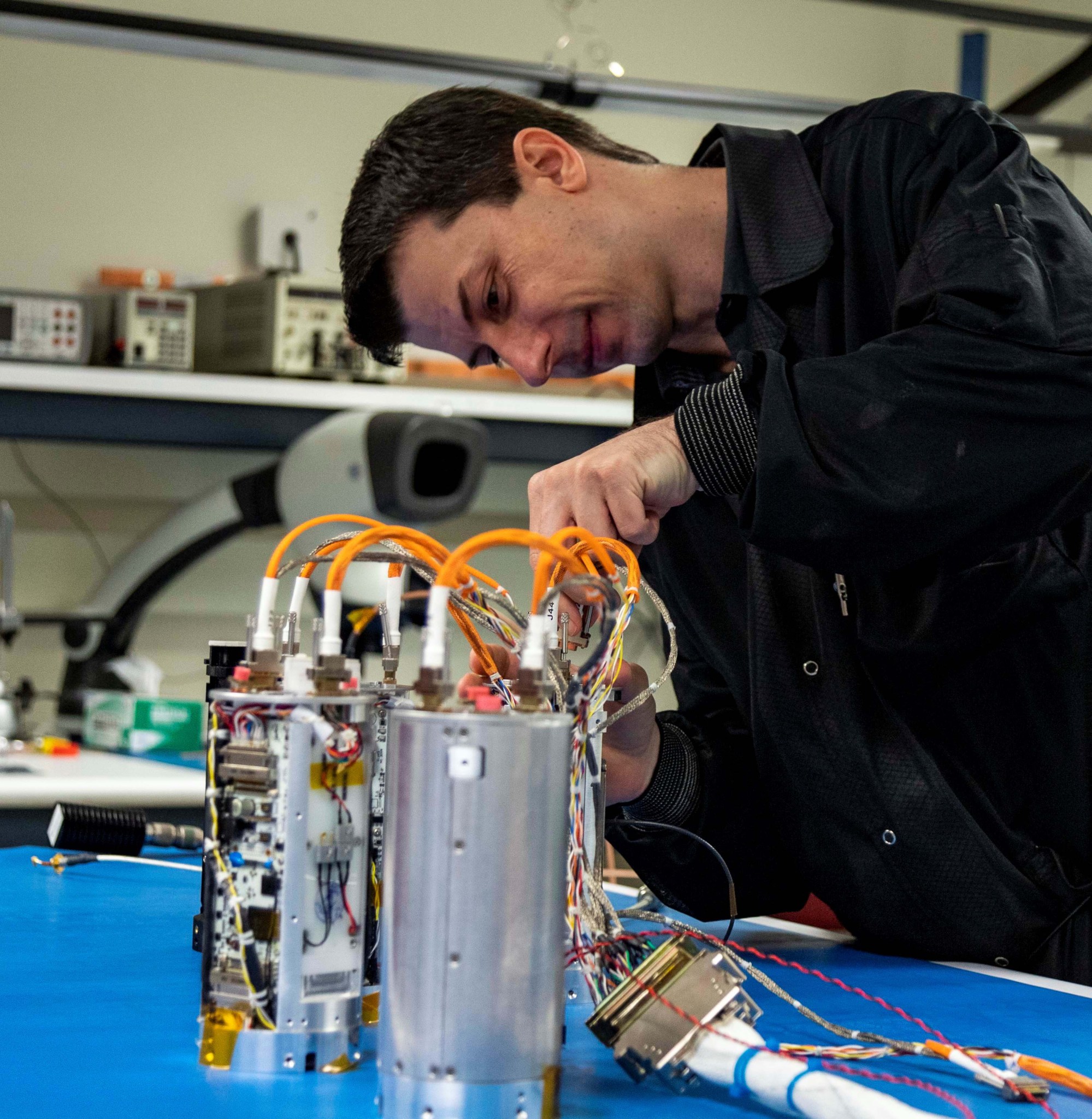 innovator: Smiling man in dark attire bent over at a blue work table, manipulating gray, shiny equipment with bundles of wires descending from the top.
