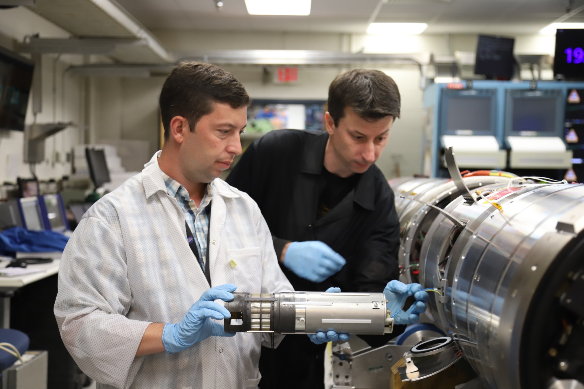Two men (one clad in an off-white lab coat, the other in dark attire) with blue gloves manipulate a small silver-gray cylinder about the size of a 1 liter water bottle into the body of a silver sounding rocket cylinder, about 18 inches wide.