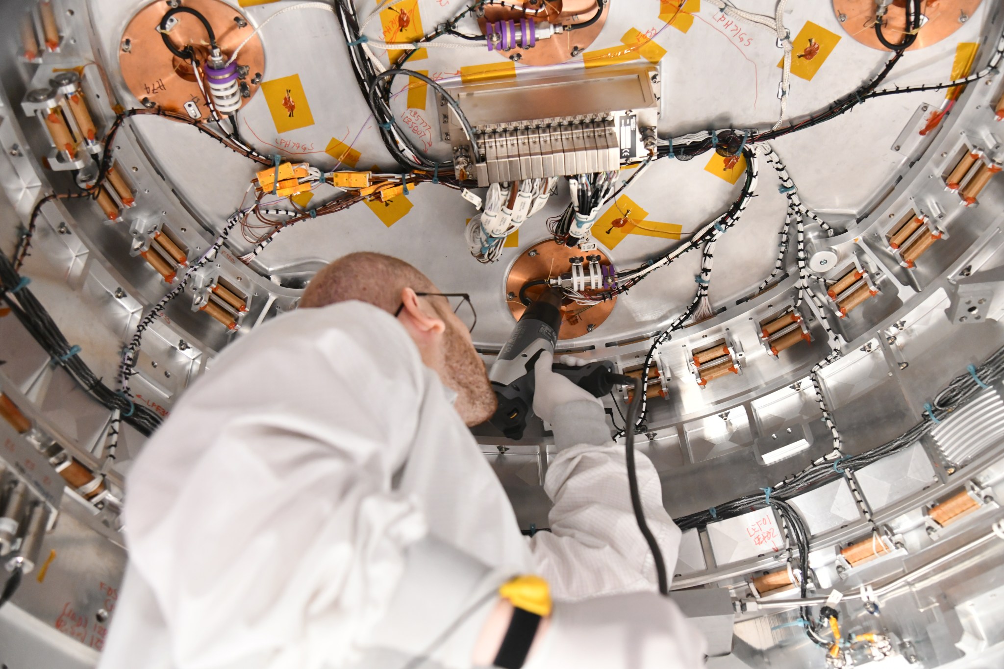 Seen from below, a person wearing a white lab coat is holding an instrument toward a panel of curving wires and technical panels.