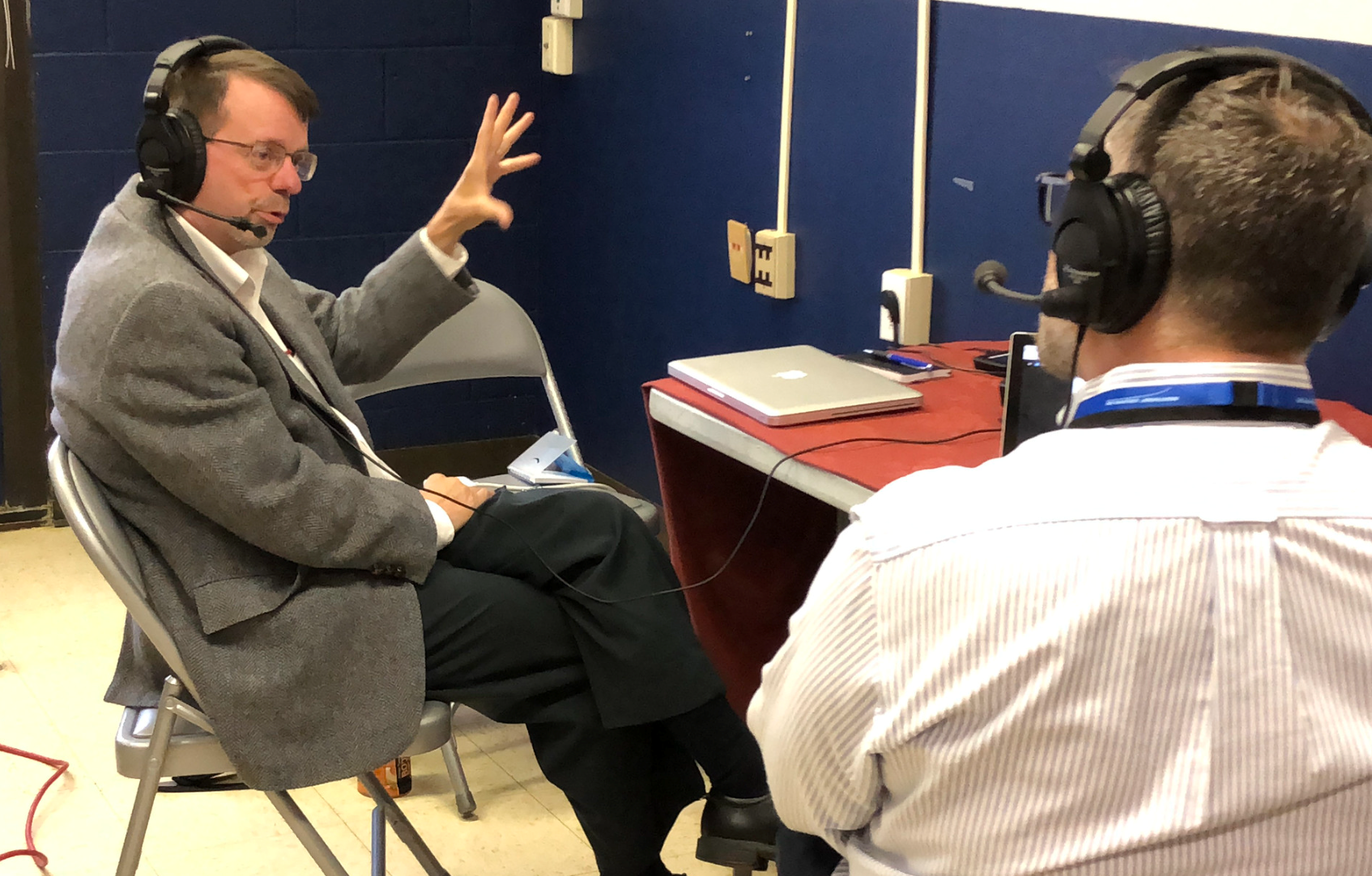Keith Koehler, a white man with short brown hair and beard, sits in a chair and gestures with one hand while doing a radio interview. He wears headphones, a taupe jacket, and dark pants.
