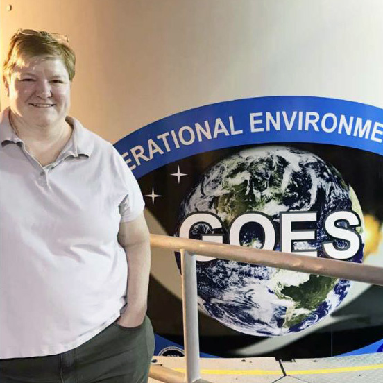 A woman stands to the left of the image wearing a light-colored polo shirt. Behind her is an image of the Earth with the letters GOES in big white letters over it.