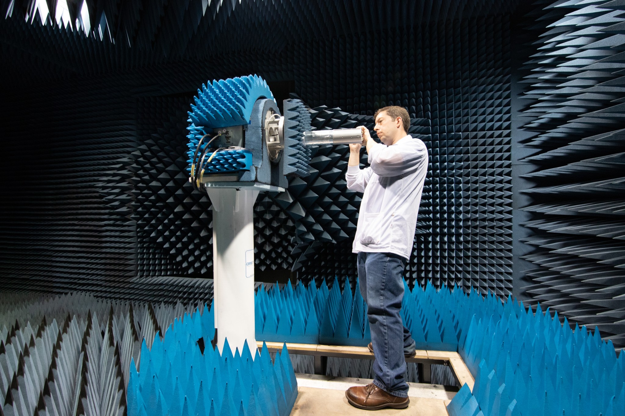 swarm payload: A man in blue jeans stands in a dimly lit room, its floor, ceiling, and walls covered in spiky, blue-gray foam pyramids. The man holds a silvery canister (about the size of a 1L water bottle) and manipulates it onto a pedestal in the center of the room.