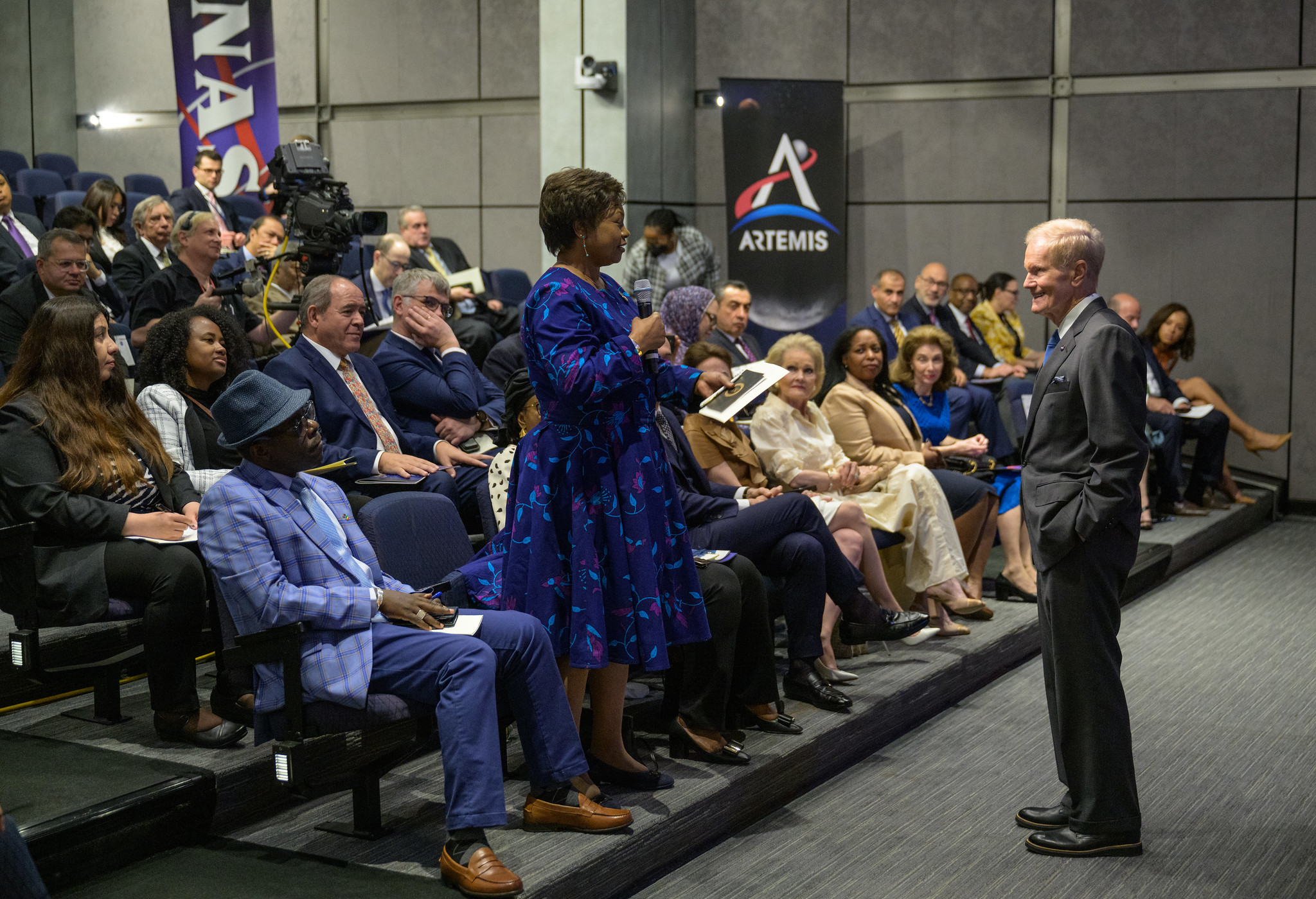In an auditorium at NASA Headquarters, NASA Administrator Bill Nelson, standing, listens to a woman who is standing and speaking into a microphone to ask a question during a U.S. Department of State Open House. Attendees of the event fill the four rows of seats visible in the image, and a banner with NASA Artemis Program logo is seen in the background.