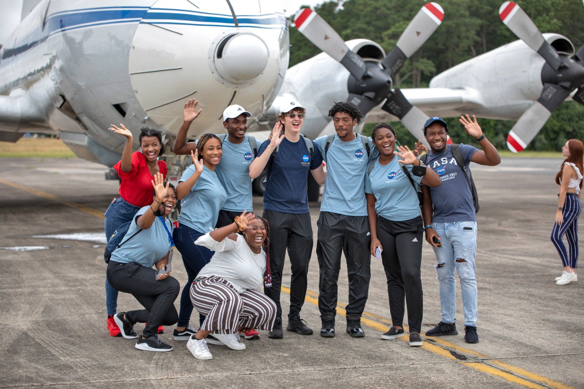 A group of nine young people wearing NASA t-shirts pose in front of the propellers and nose of an airplane on a runway.