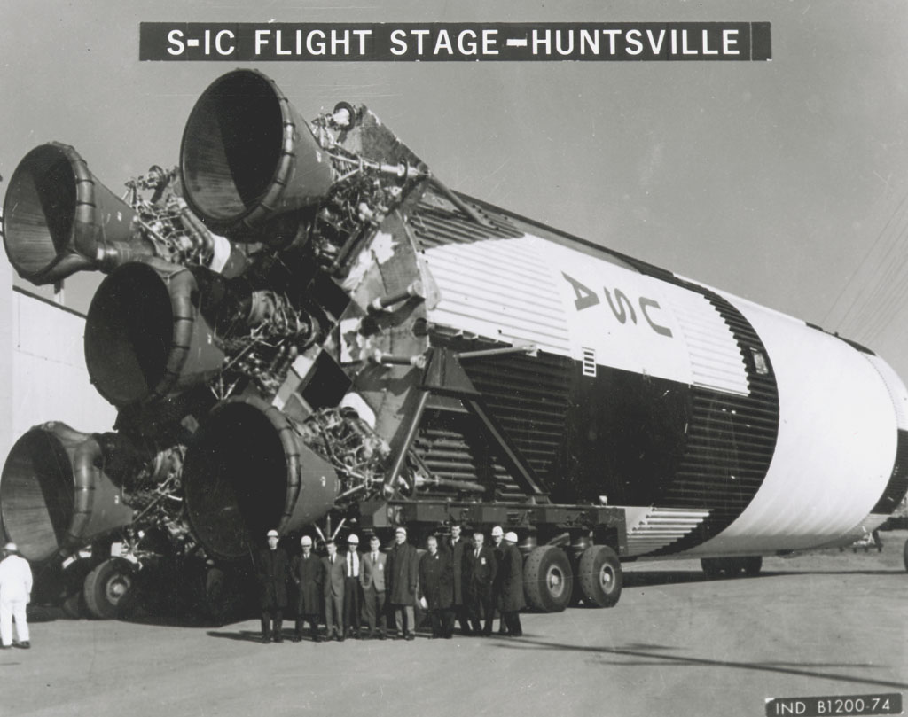 A Saturn V lies on its side as a group of men stand in front of it in this black and white photo. A label of S-IC Flight Stage - Huntsville is at the top.