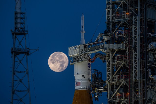 A close-up view of NASA's Orion spacecraft atop the Space Launch System at Launch Complex 39B with a full Moon in the background.
