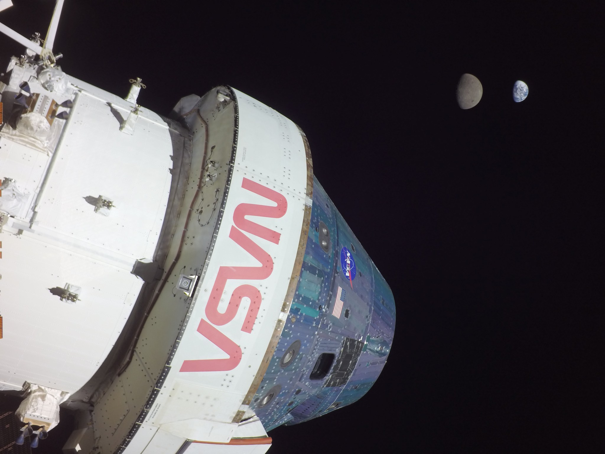 The Earth and Moon appear side by side off in the distance while the Orion crew module is in the foreground.