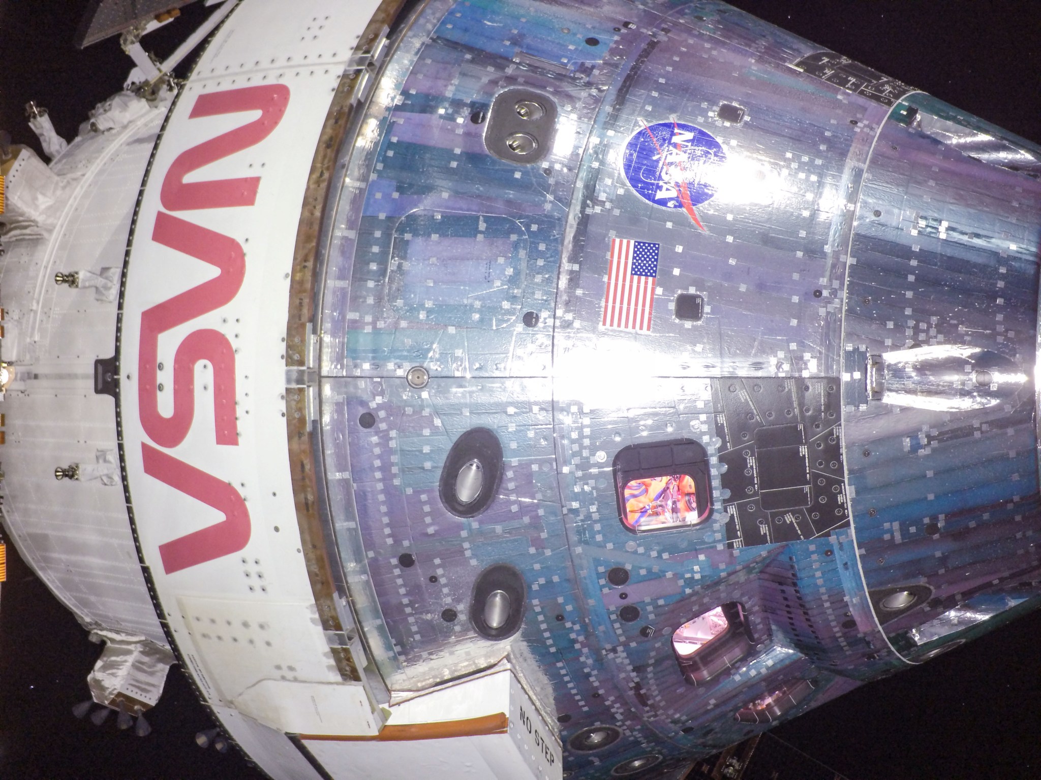 A close up view of the Orion in space. The orange spacesuit on Commander Moonikin Campos can be seen through one of the crew module's windows. The American flag on the body of the crew module and red NASA lettering can also be seen.