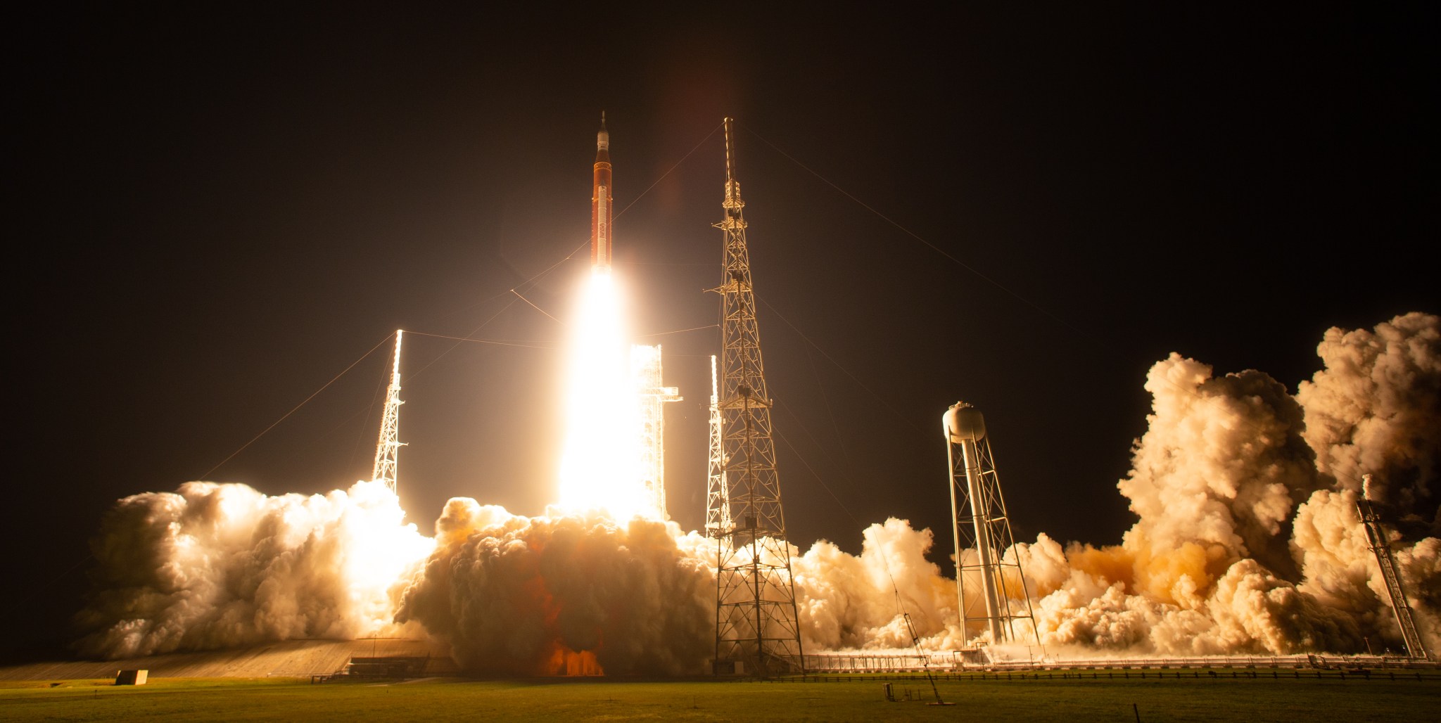 The Space Launch System rocket ascends above the mobile launcher and lights up the night sky, with smoke from liftoff billowing across the launch pad area.