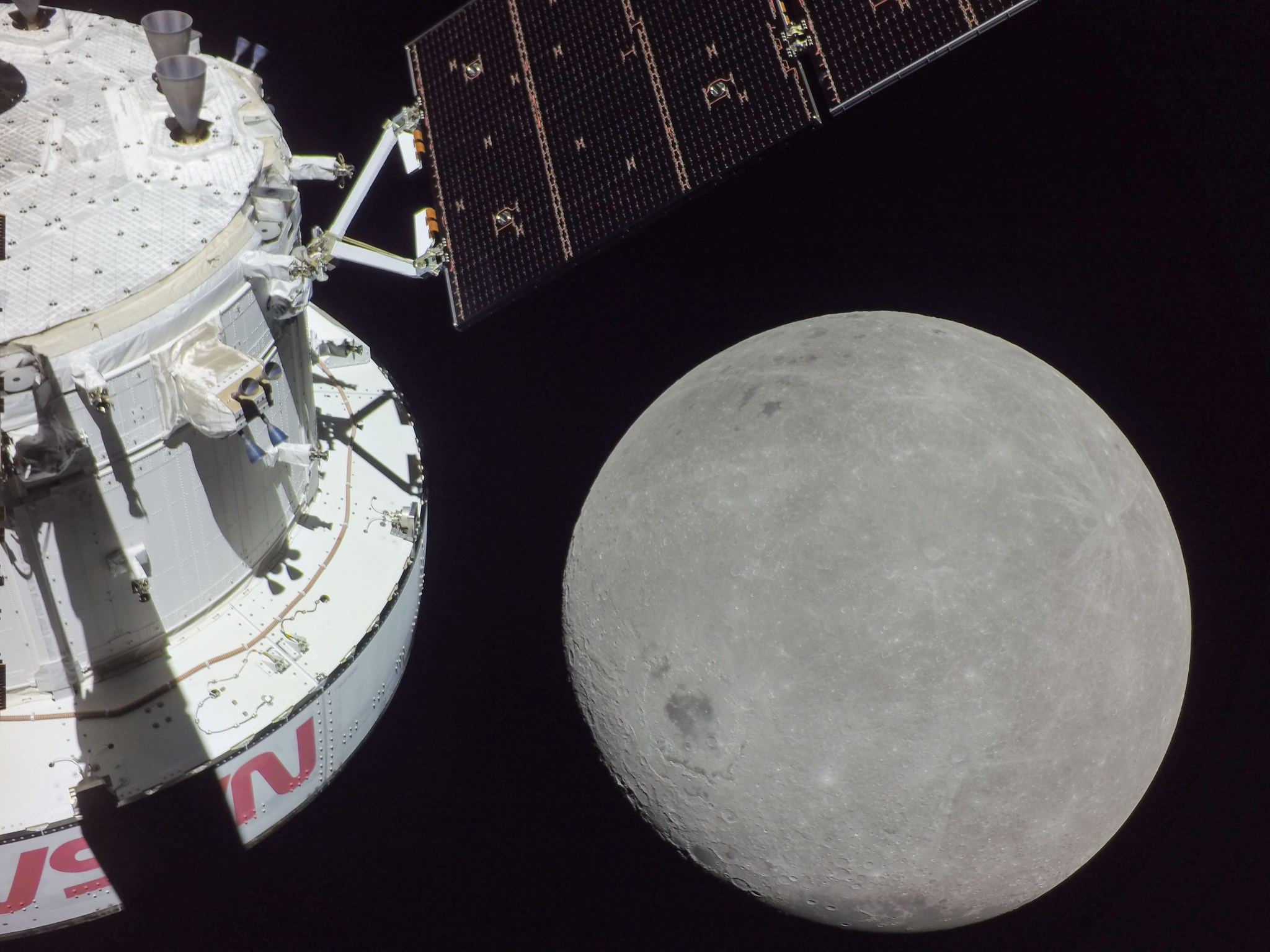 The grey, circular Moon is seen as the Orion spacecraft approaches it. The back end of the service module, including a solar array wing, and red NASA lettering can be seen. 