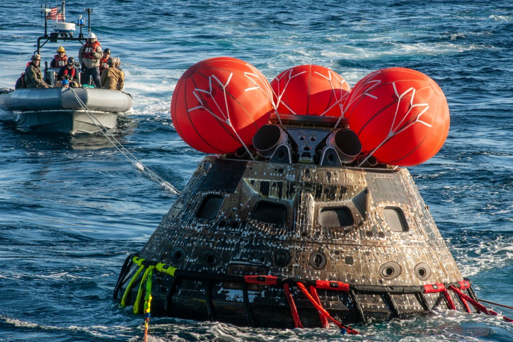 The Orion crew module is seen in the Pacific Ocean, with several red, circular airbags atop it can be seen, while a small boat with recovery personnel approaches the capsule.
