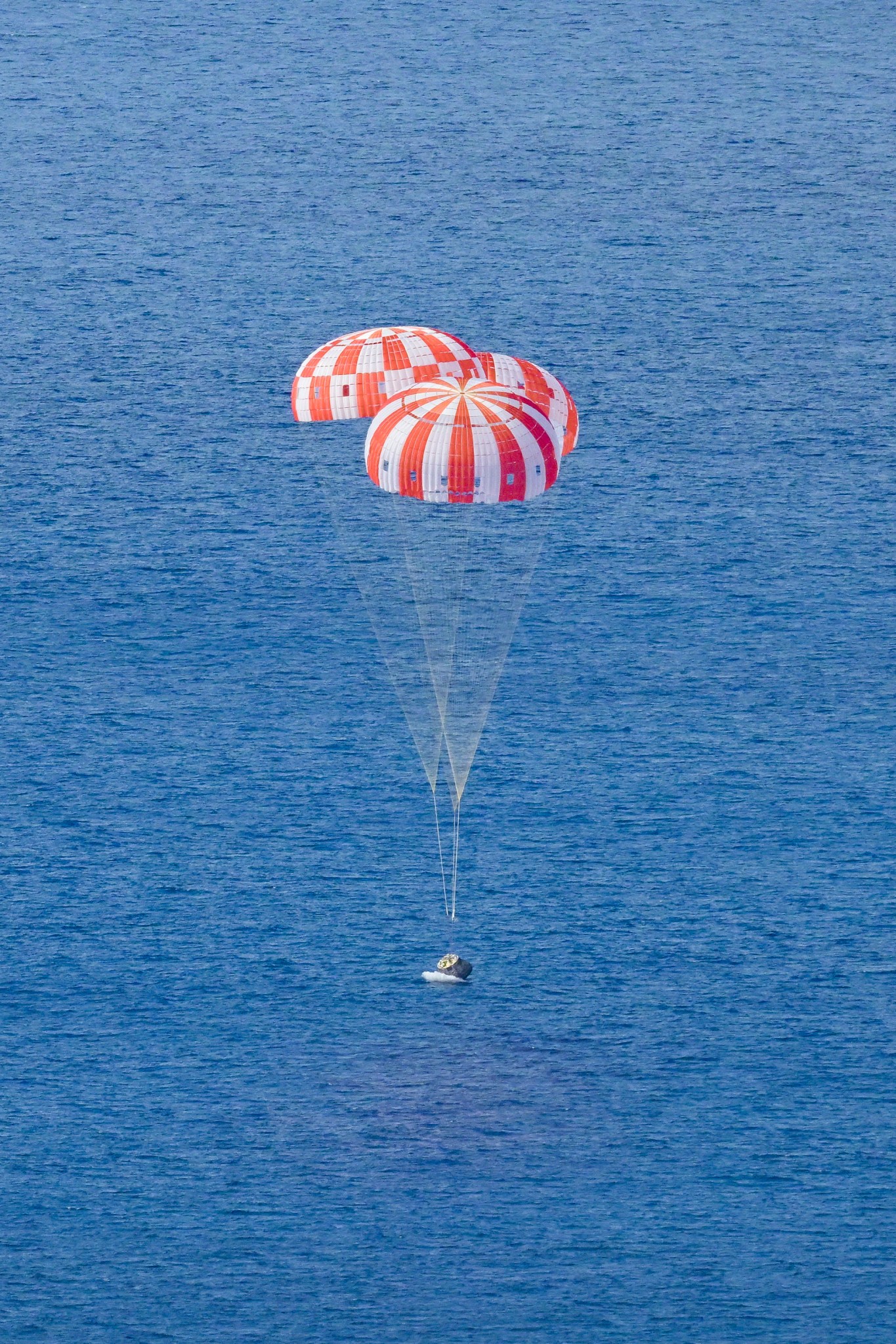 The Orion crew module, with its orange and white main parachutes fully expanded, dips at an angle into the Pacific Ocean at the moment of splashdown. 